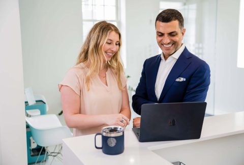 Two professionals in a modern clinic setting collaborate and review something on a laptop, highlighting teamwork, communication, and training often found in high-performing chiropractic teams. The scene is bright, wide, and inviting, conveying operational excellence and a seamless patient experience.